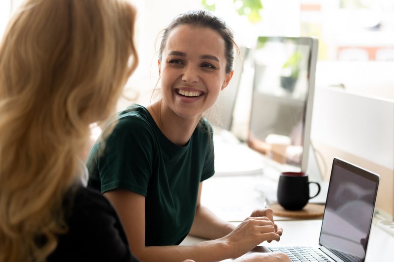 Zwei Frauen lachen bei der Arbeit vor einem Laptop. 