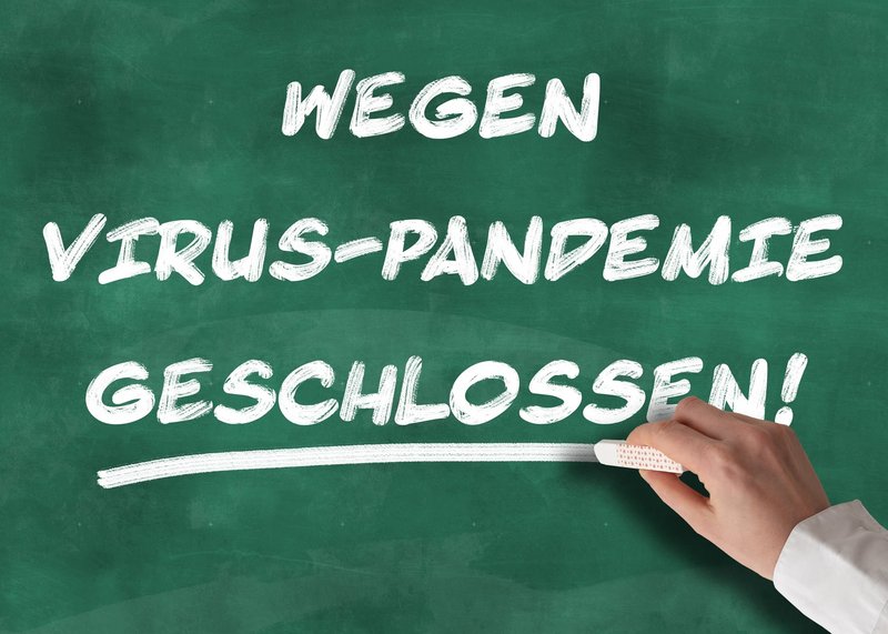 Hand schreibt mit Kreide "Wegen Virus-Pandemie geschlossen!" an eine Tafel.