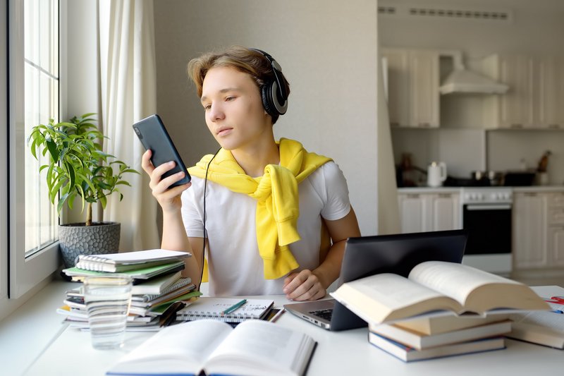 Schüler sitzt mit Kopfhörern und Handy vor einem Schreibtisch mit geöffneten Büchern und einem Laptop. 