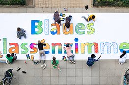 Das Blaue Klassenzimmer Kinder bemalen ein großes Banner, am Boden liegend, mit der Aufschrift "Blaues Klassenzimmer"