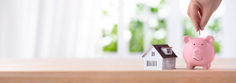Hand tut Geld in ein Sparschwein neben dem ein kleinen Haus steht. 