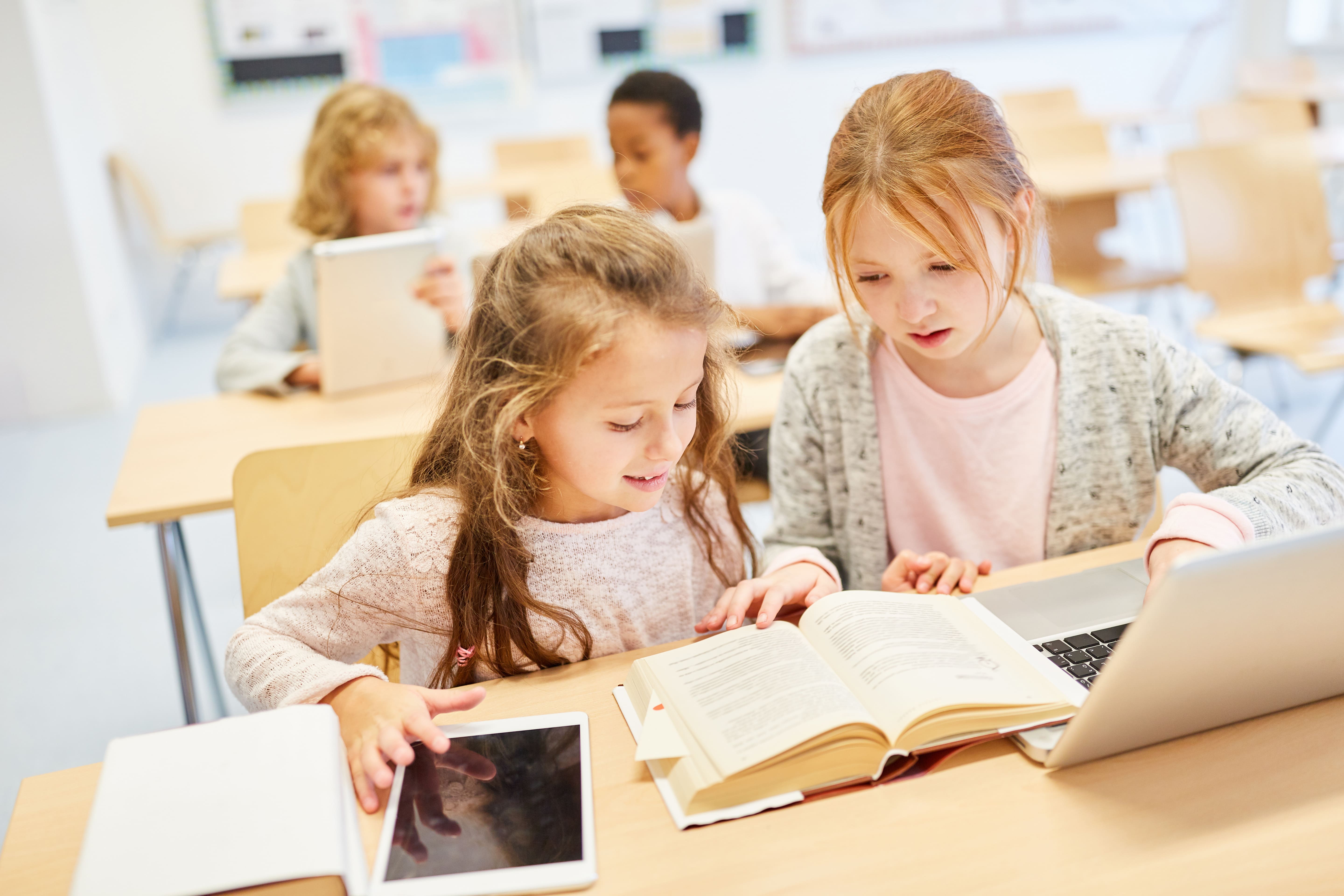 Zwei Mädchen im Klassenzimmer mit aufgeschlagenem Buch und Tablet