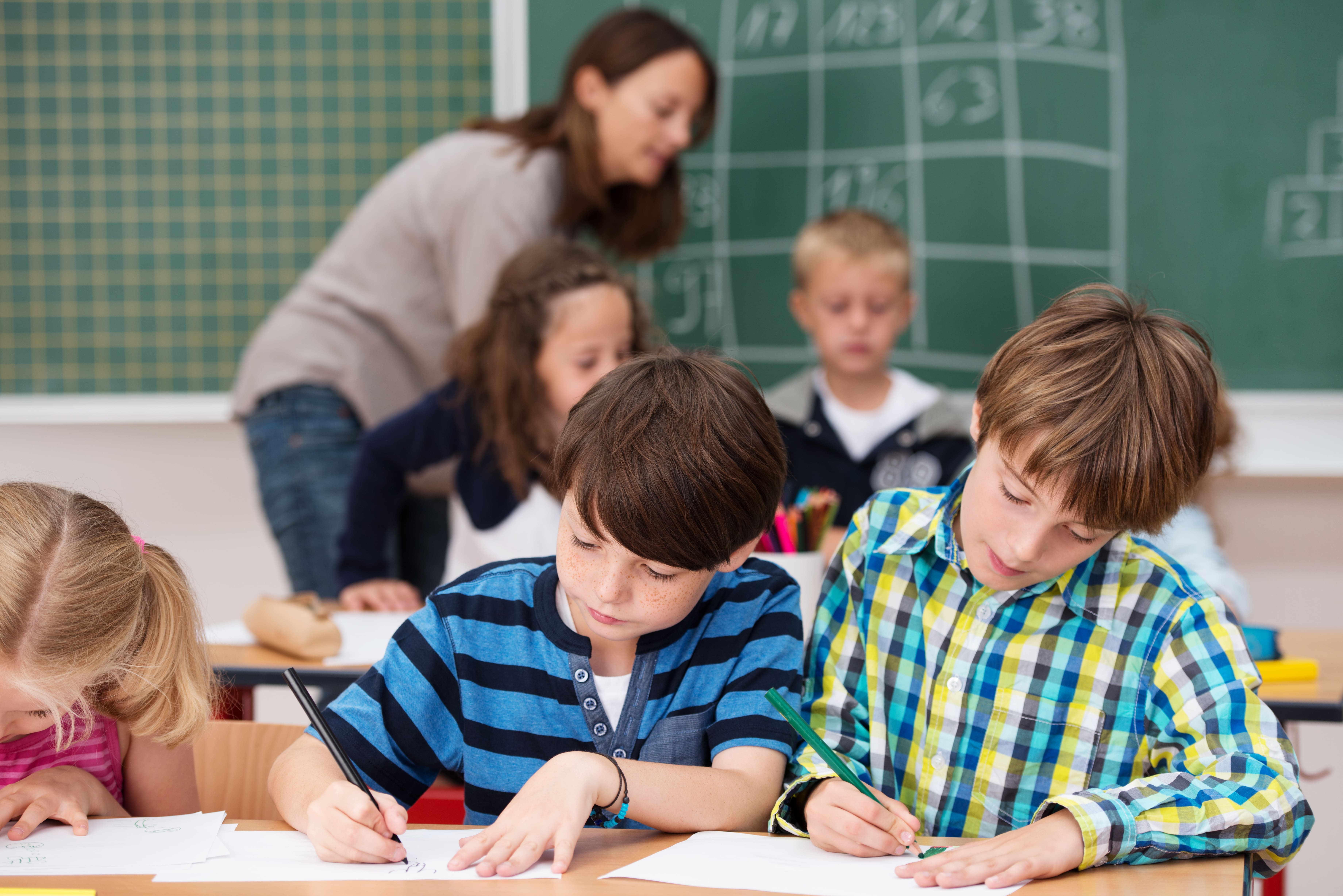 Lehrkraft als Lernbegleitung im Hintergrund, im Vordergrund schreibende Schülerinnen und Schüler