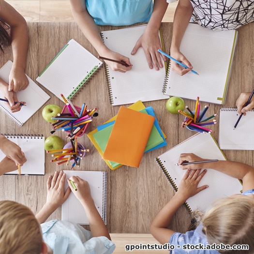 Viele Schreibblöcke liegen auf einem Tisch und man sieht die Hände von Kindern, die schreiben.