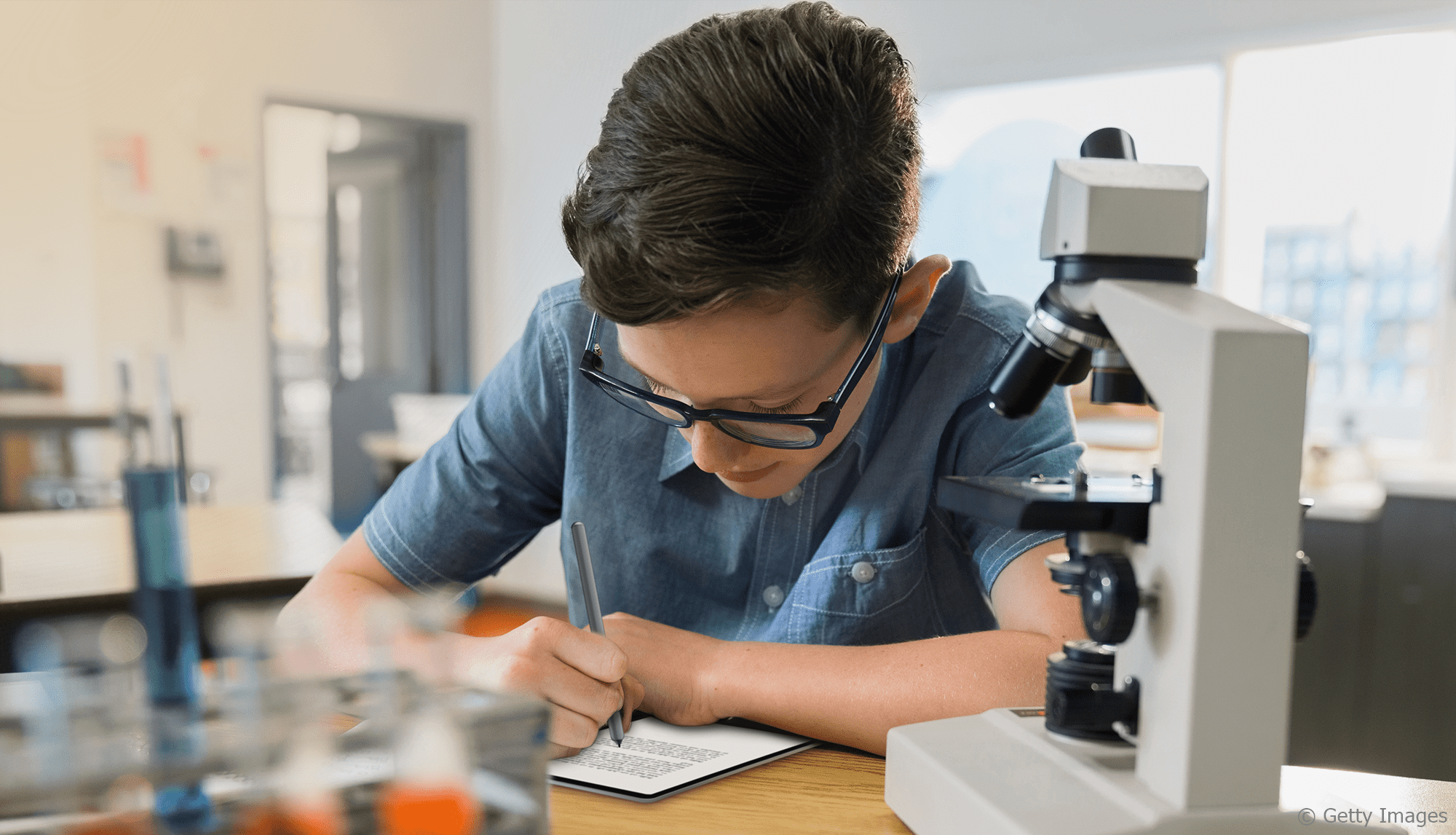Ein Schüler sitzt neben einem Mikroskopt und schreibt etwas mit einem Stift auf ein Tablet